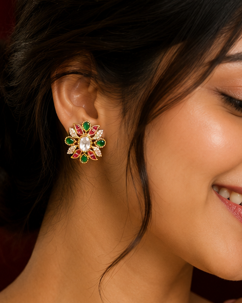 A woman wearing a cream-colored saree and a matching cream blouse, shown in a close-up portrait. She is wearing a prominent gold-toned floral stud earring decorated with red and green stones.