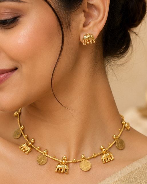 A woman wearing a gold-toned elephant charm necklace and matching stud earrings, styled with a beige saree in a warm, elegant indoor setting.