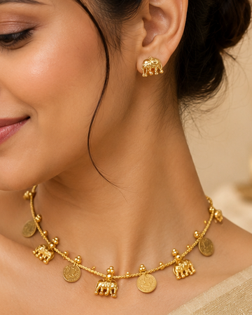 A woman wearing a gold-toned elephant charm necklace and matching stud earrings, styled with a beige saree in a warm, elegant indoor setting.