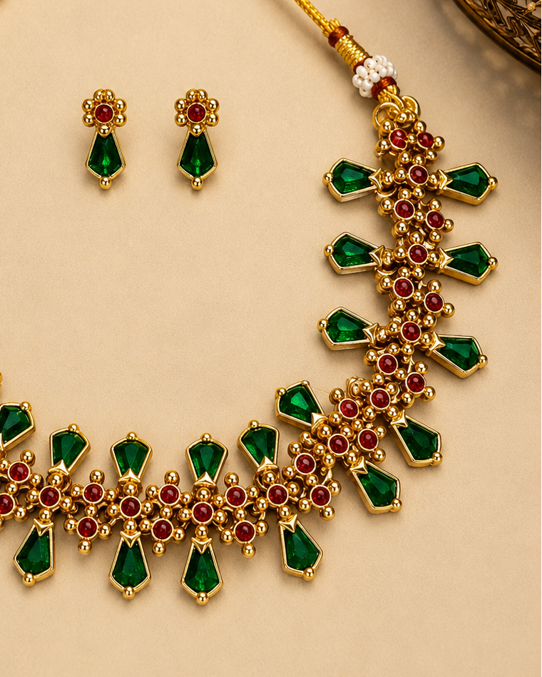 Gold-plated statement necklace and matching stud earrings set with green teardrop-cut stones and red round stones, arranged on a beige background with decorative dried flowers and gold fabric.