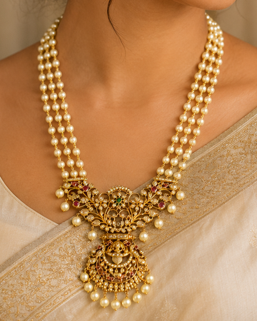 A woman wearing an ornate gold and pearl traditional necklace with matching dangling earrings, styled with a cream-colored sari in a warm, elegant indoor setting.