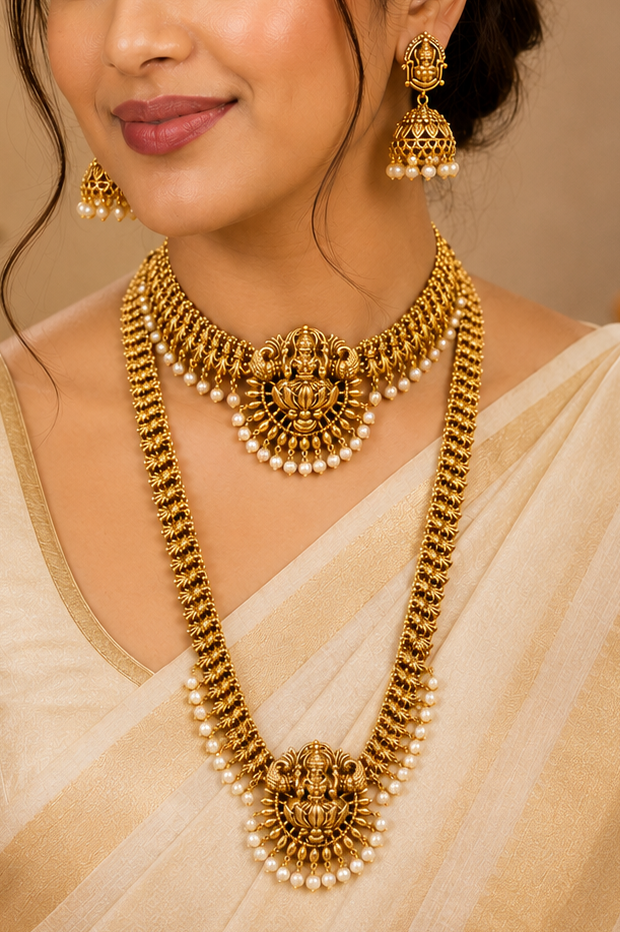 A woman wearing an ornate gold-toned traditional necklace with matching earrings and a cream-colored saree, photographed in a warm indoor setting with soft lighting.