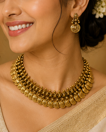 A woman wearing a traditional Indian outfit with a beige saree and a prominent gold multi-strand collar necklace, paired with matching gold earrings.