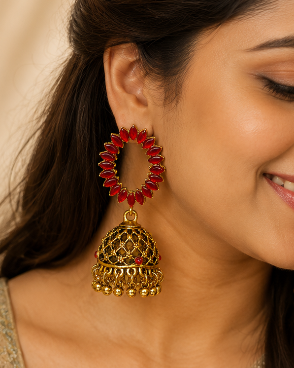 A woman wearing large traditional Indian jhumka earrings with red circular beadwork and gold-toned bell-shaped dangling charms, shown in a close-up portrait.
