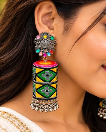A close-up lifestyle shot of a woman wearing ornate traditional Indian jhumka earrings. The earrings feature a silver-toned metal frame adorned with colorful rhinestones, a pink and yellow top, and a cylindrical tassel decorated with green and yellow geometric patterns and silver bell charms.