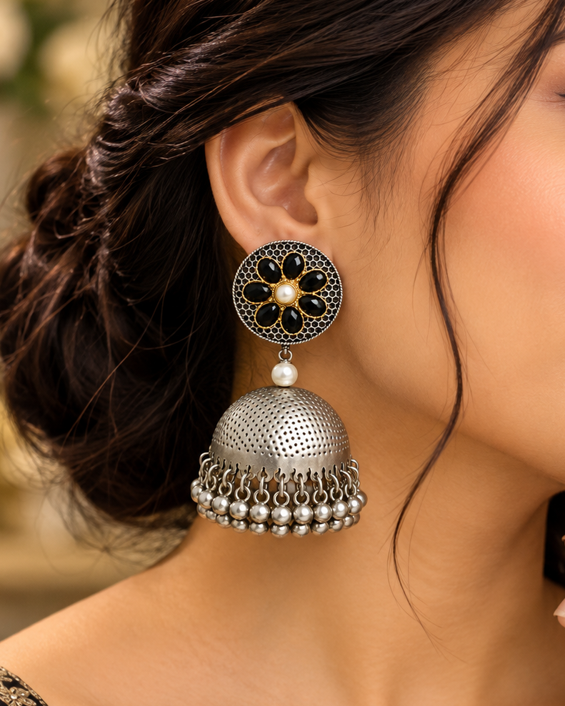 A close-up of a woman wearing a large traditional Indian jhumka earring. The earring features a black and gold floral filigree top, a small white pearl, and a silver-toned bell-shaped dangle with hanging silver beads.