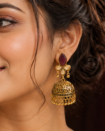 A woman wearing ornate gold jhumka earrings with dark red oval stones, styled with a cream saree, in a warm, elegant indoor setting.