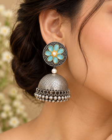 A close-up of a woman wearing ornate silver jhumka earrings with turquoise blue floral details and small pearl-like accents, against a soft beige background.