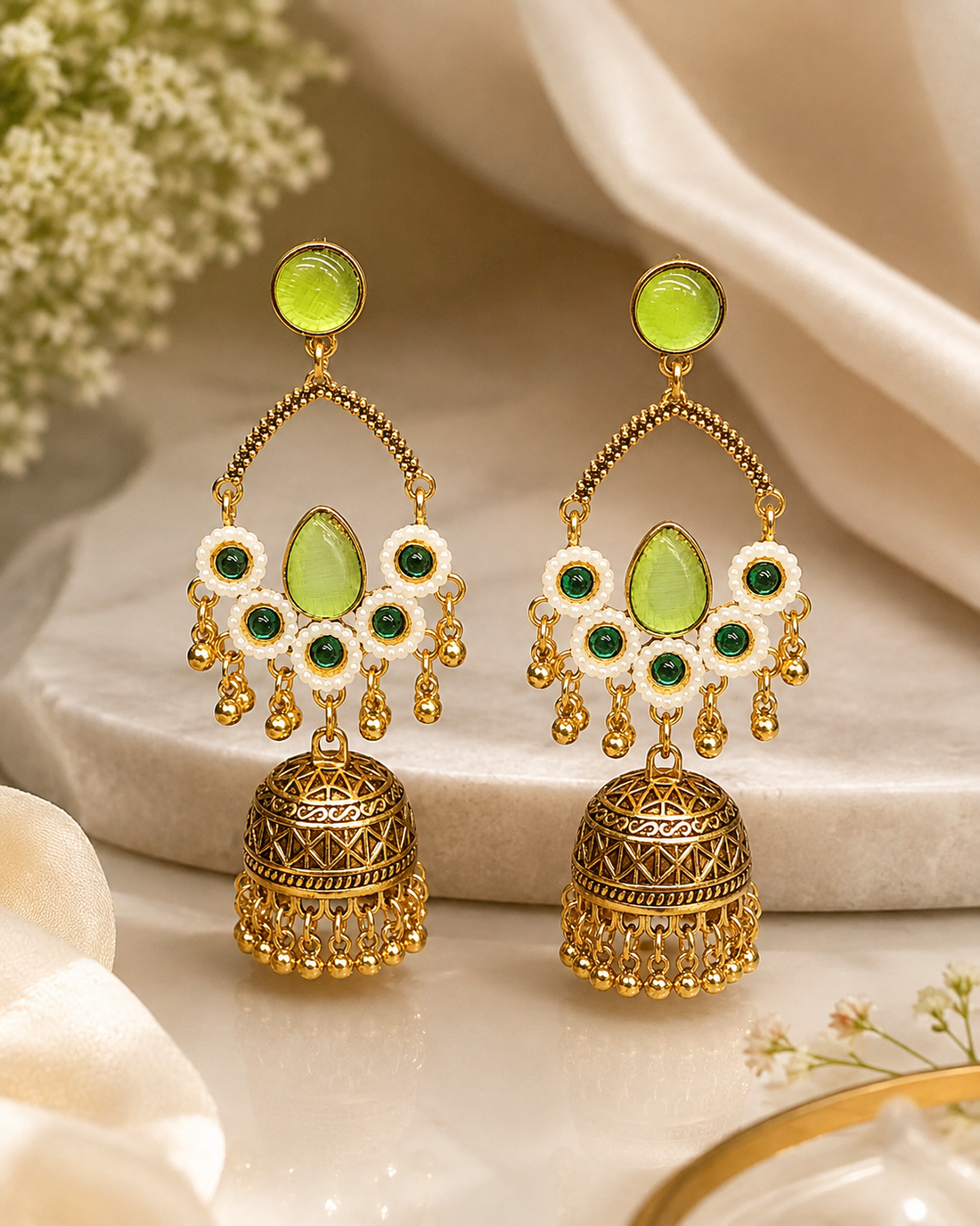 A woman in profile wearing ornate gold-toned jhumka earrings with light green stones and white enamel details, paired with a cream-colored sari.