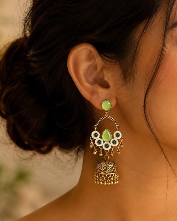 A woman in profile wearing ornate gold-toned jhumka earrings with light green stones and white enamel details, paired with a cream-colored sari.