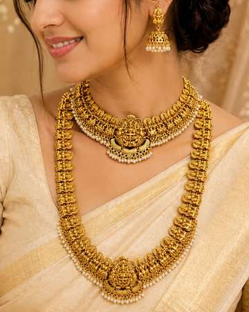 A woman wearing an ornate traditional gold necklace with matching earrings and a cream-colored saree, shown in a warm, elegant indoor setting.