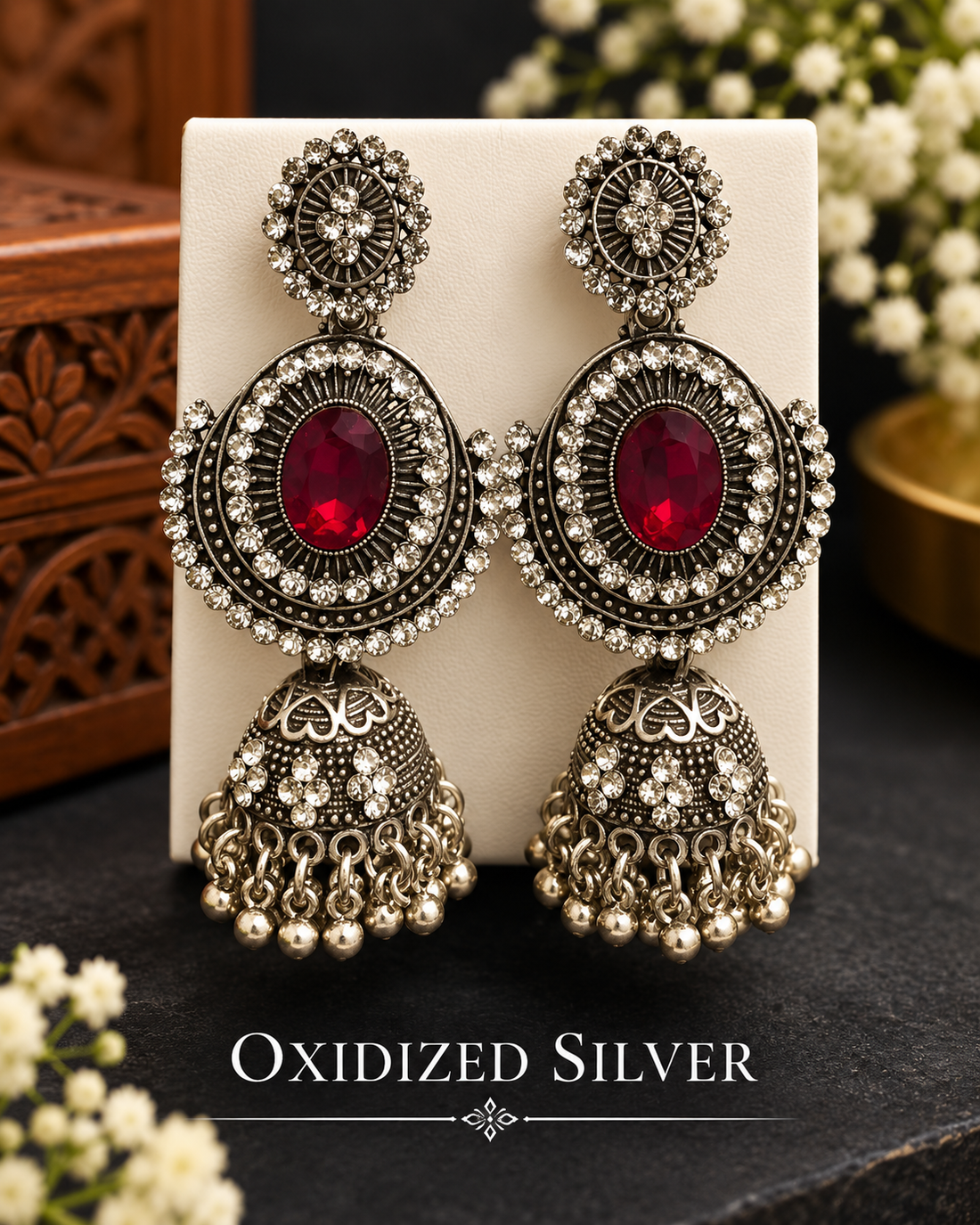 A close-up lifestyle shot of a woman wearing a large, ornate silver-toned dangle earring with a central red gemstone. The earring features intricate silver filigree, a round silver disc, and a dangling bell-shaped charm adorned with small silver beads.
