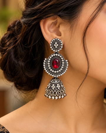 A close-up lifestyle shot of a woman wearing a large, ornate silver-toned dangle earring with a central red gemstone. The earring features intricate silver filigree, a round silver disc, and a dangling bell-shaped charm adorned with small silver beads.