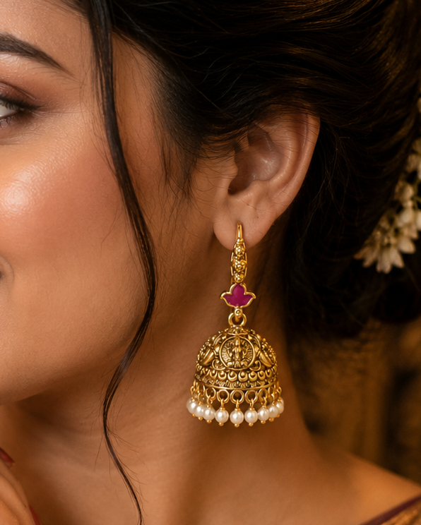 A woman in a traditional Indian saree wearing an ornate gold jhumka earring with a pink stone and white pearl accents, set against a warm, softly blurred background.