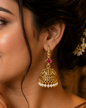 A woman in a traditional Indian saree wearing an ornate gold jhumka earring with a pink stone and white pearl accents, set against a warm, softly blurred background.