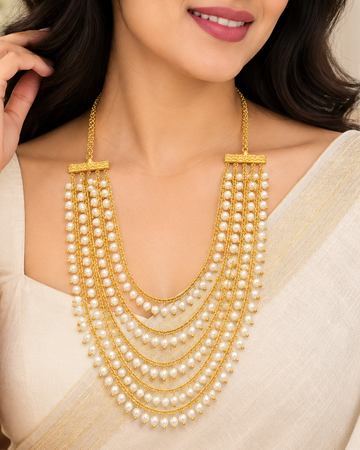 A woman wearing a multi-strand gold and pearl necklace with a traditional Indian outfit, shown in a warm indoor setting.