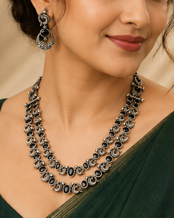 A woman wearing a dark green saree with a gold border and a matching double-strand statement necklace with silver-toned stones, set against a warm beige background.
