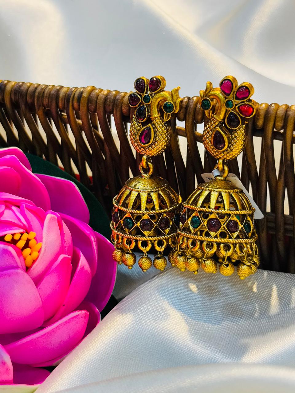 Gold earrings with red and green gemstones on a white fabric background with a pink flower.