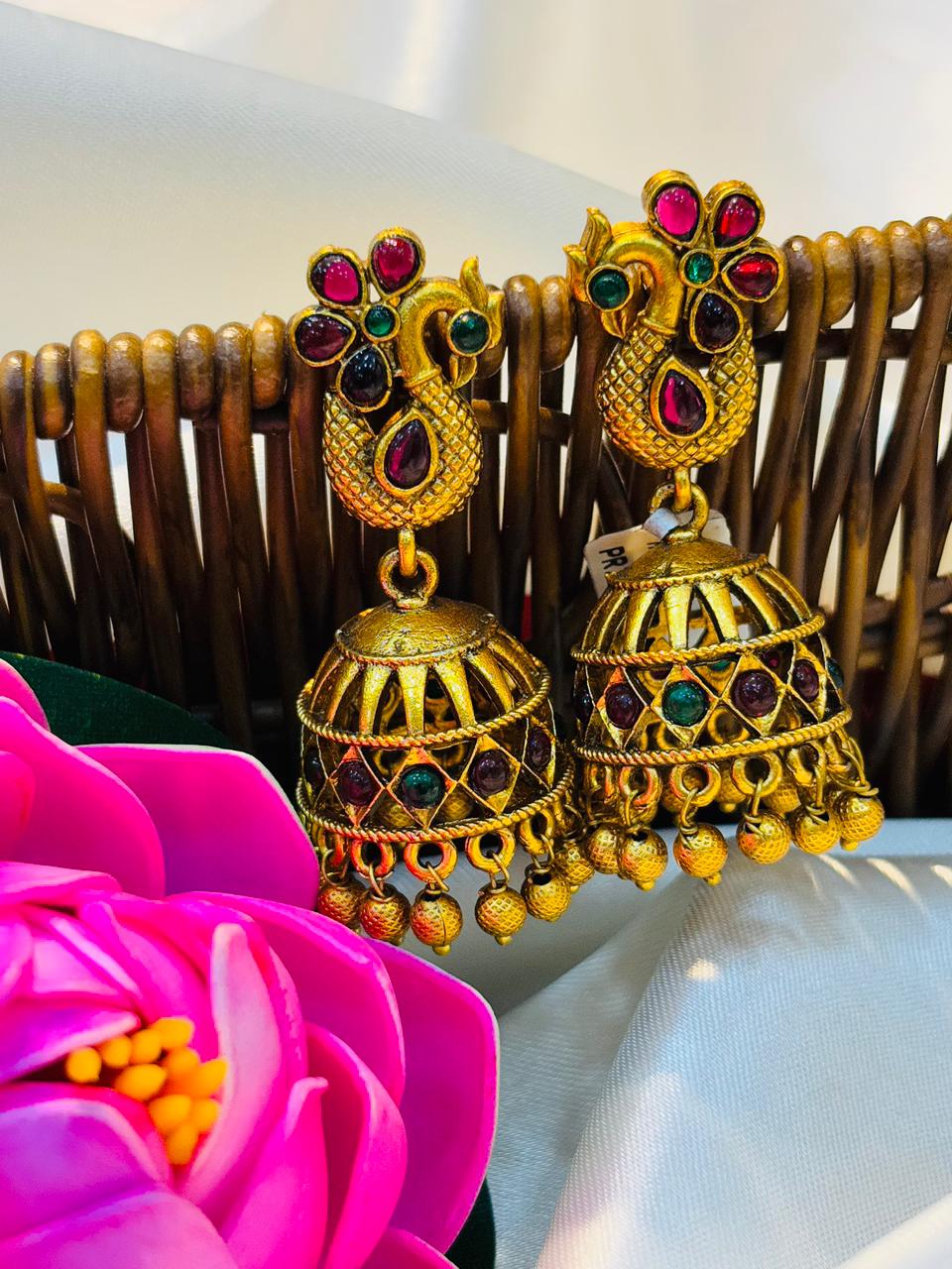 Gold jhumka earrings with colorful stones on a white surface with a pink flower.