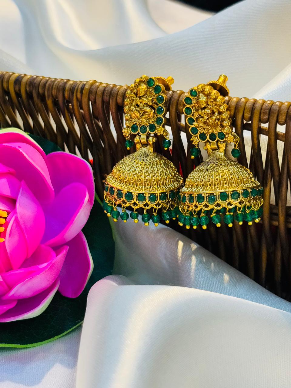 Gold and green earrings on a white surface with a pink flower and wicker basket in the background.