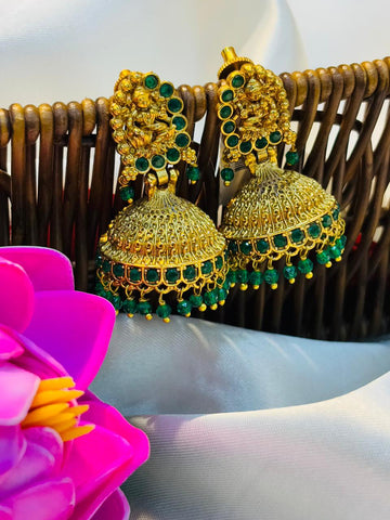 Gold and green earrings on a white surface with a pink flower and wicker basket in the background.