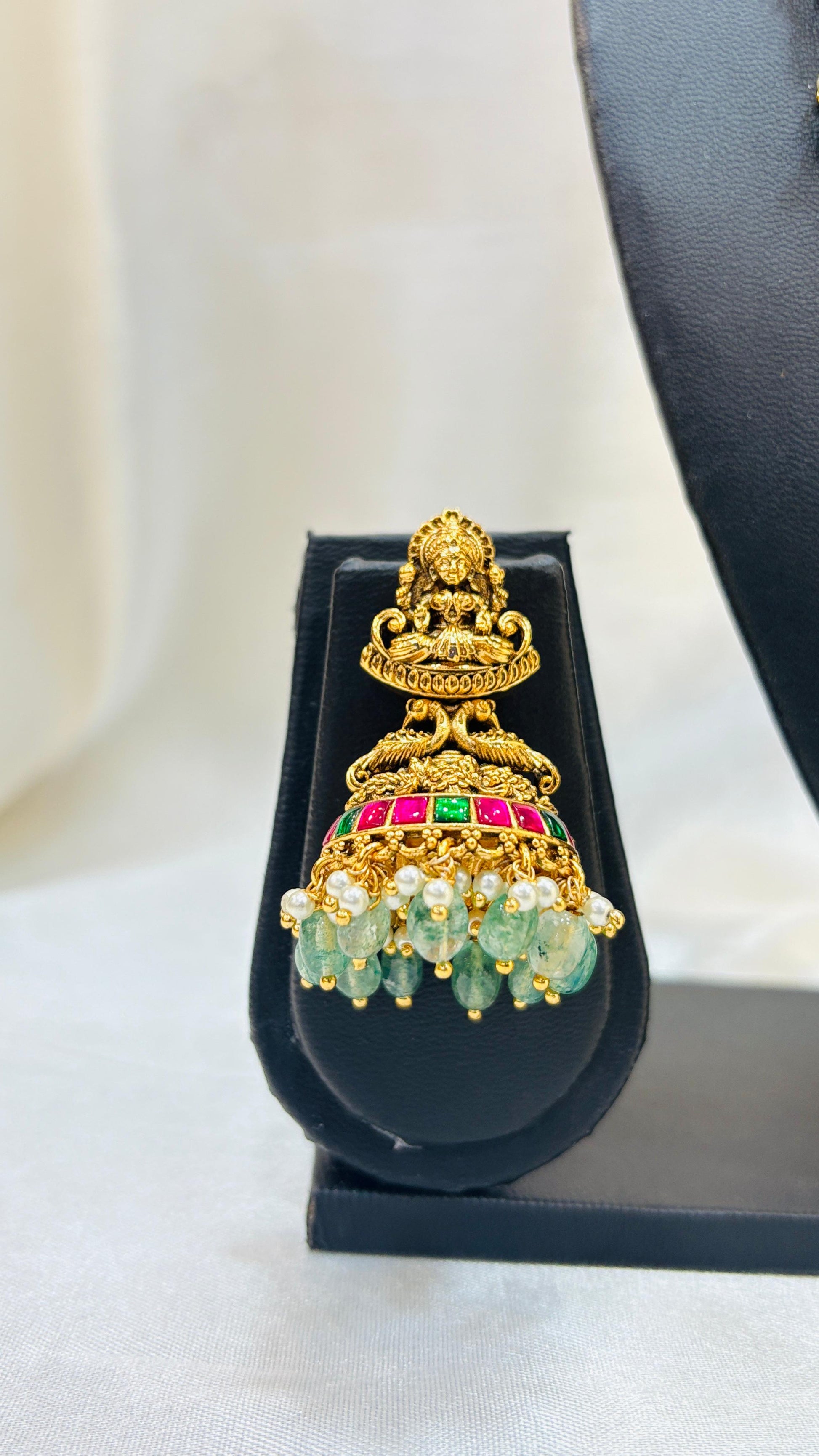 Gold earring with green and red stones on a black stand against a white background