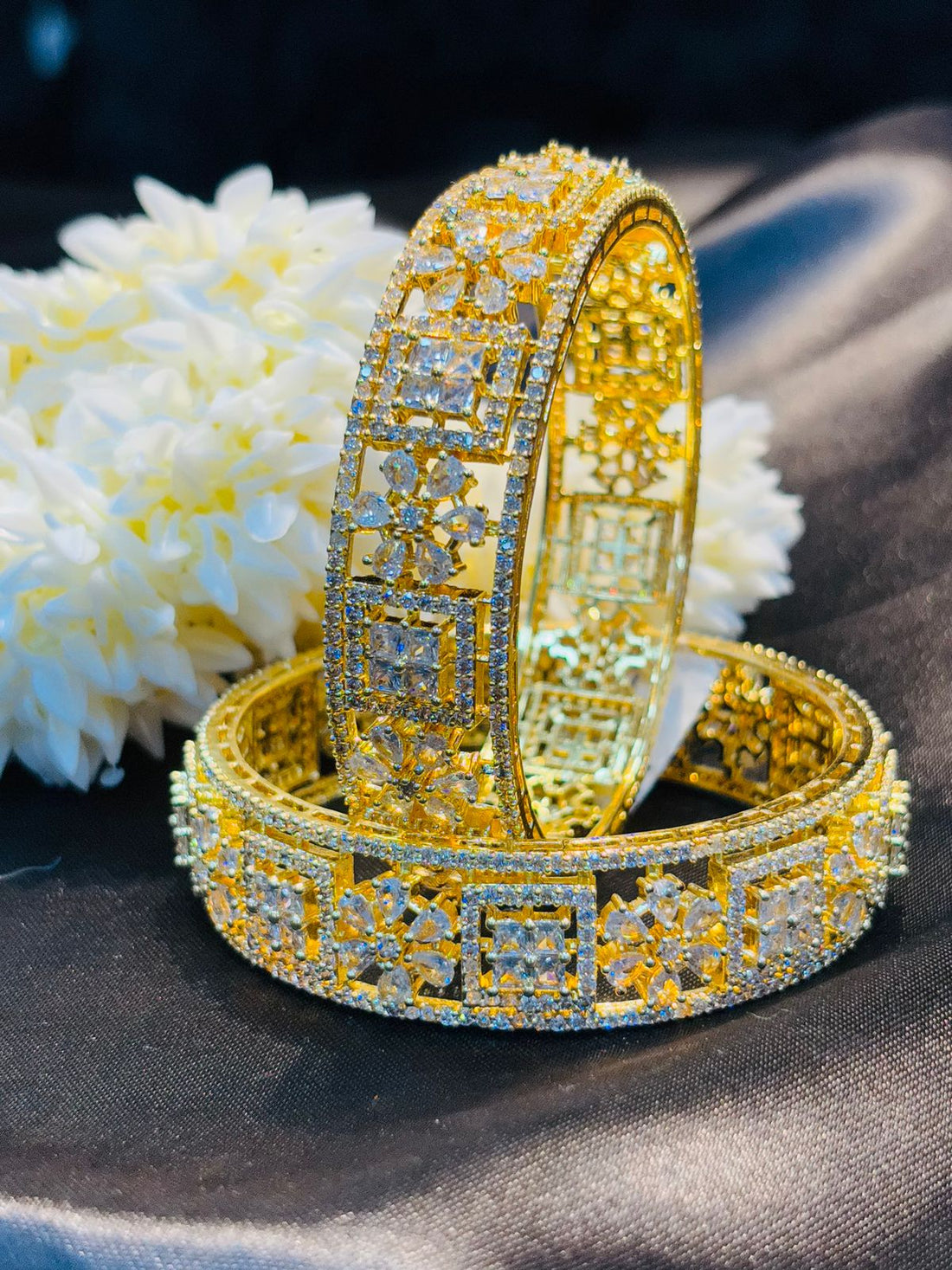 Two gold bracelets with intricate designs on a dark fabric background with white flowers.