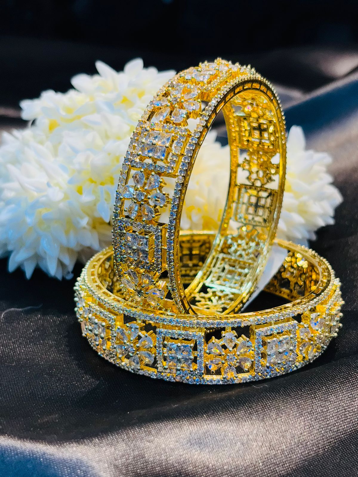 Two gold bracelets with intricate designs on a dark fabric background with white flowers.