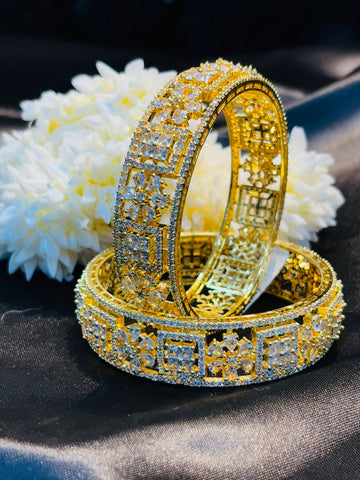 Two gold bracelets with intricate designs on a dark fabric background with white flowers.