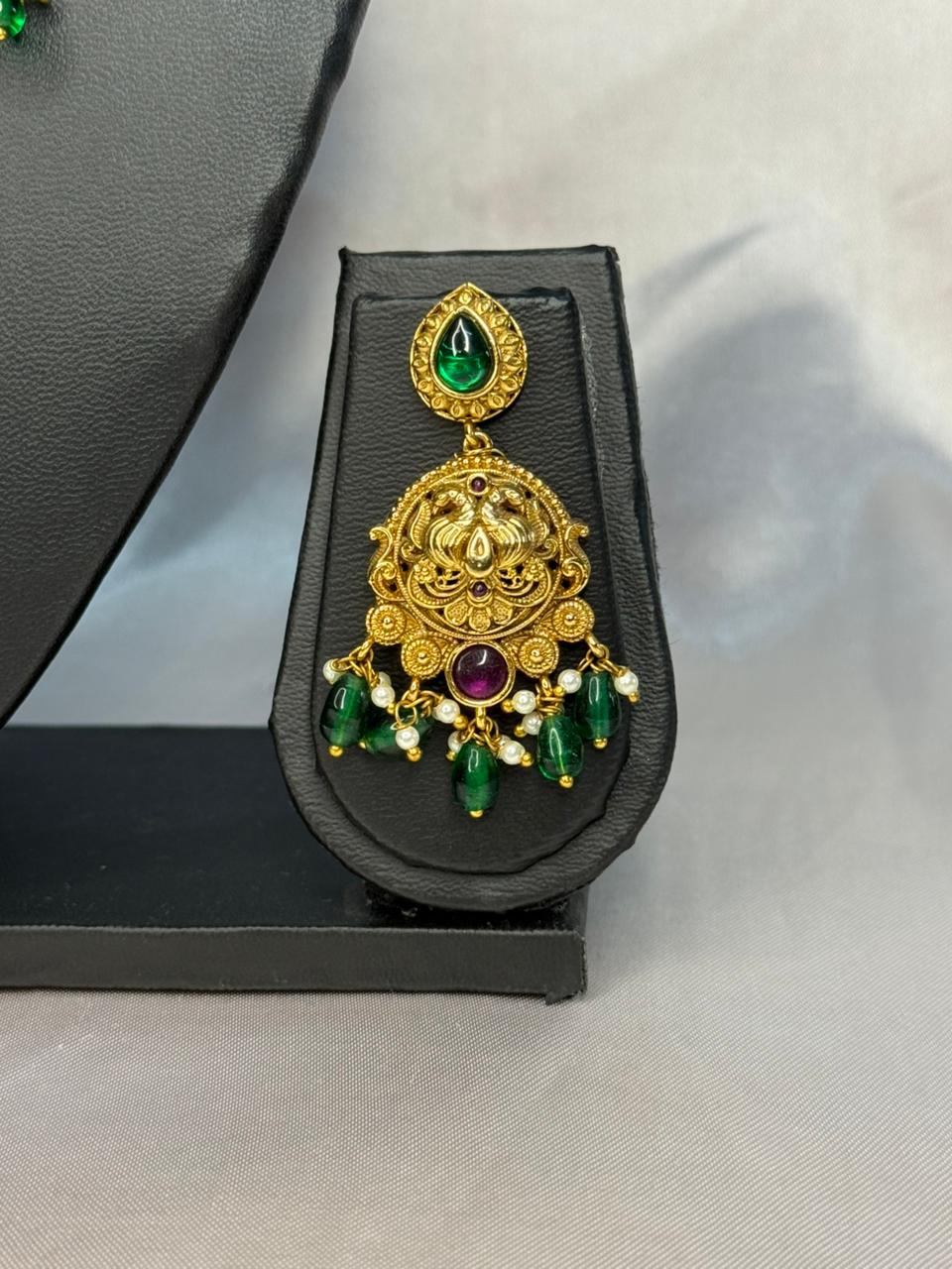 Ornate gold earrings with green and red gemstones on a black stand.