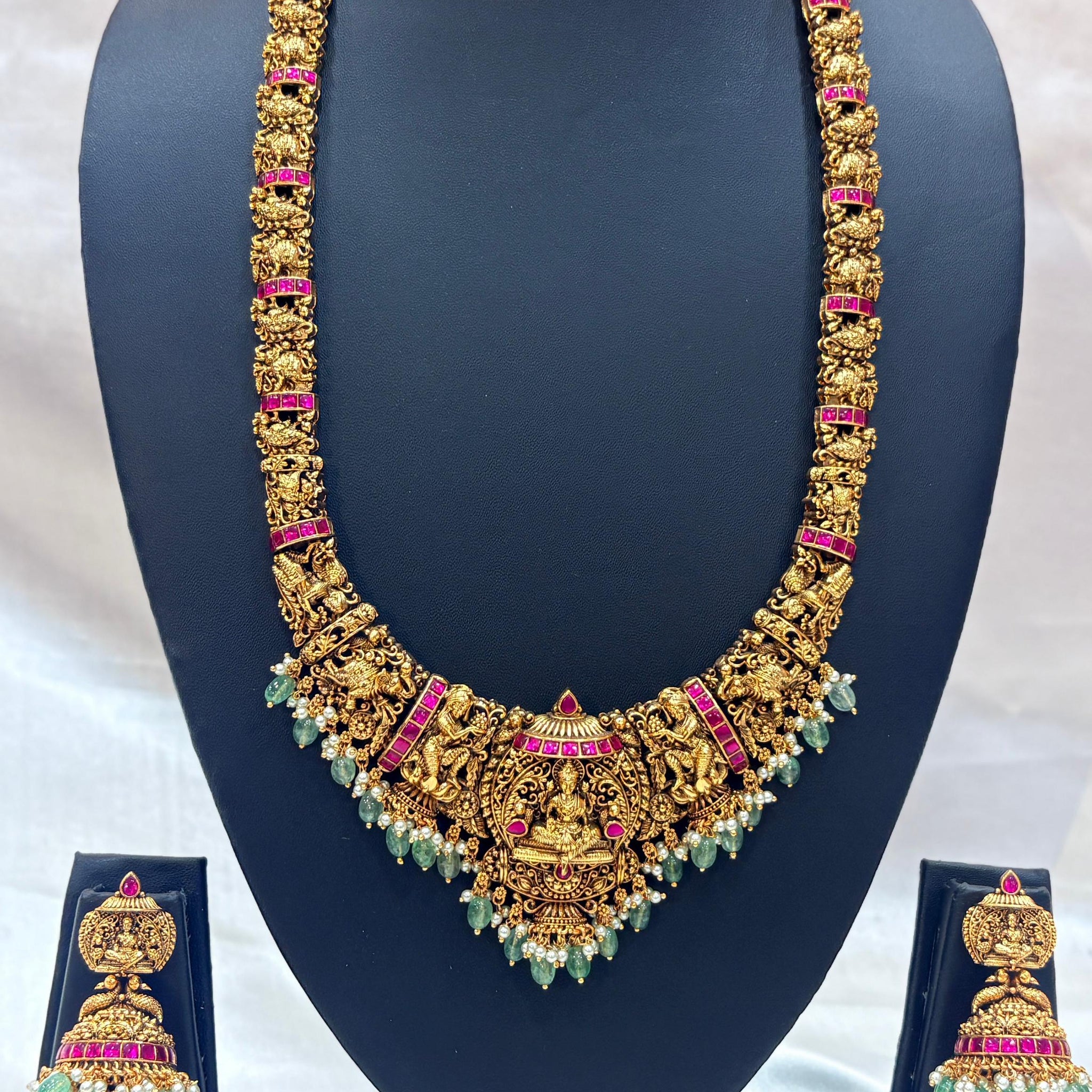 Ornate gold necklace with green and pink stones on a black stand