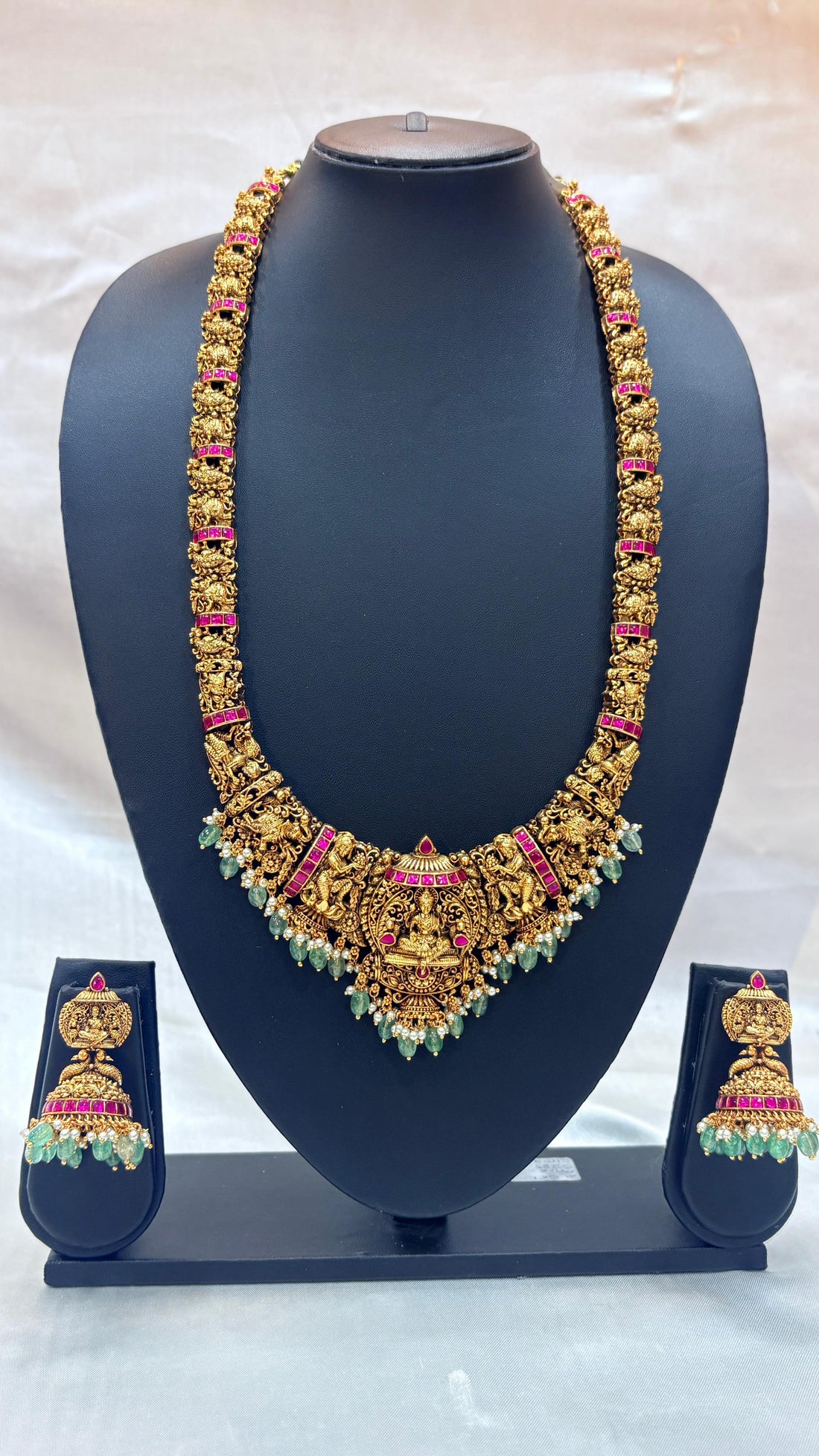 Ornate gold necklace with green and pink stones on a black stand