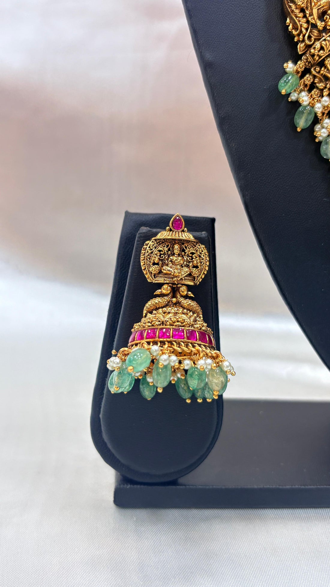 Ornate gold necklace with green and pink stones on a black stand